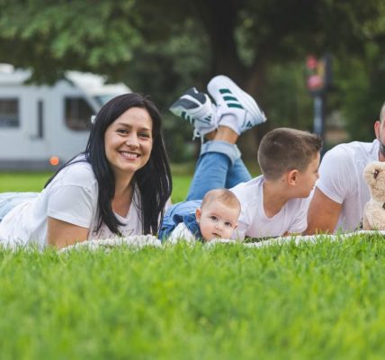 Happy family lying on grass with RV in the background, enjoying a sunny outdoor picnic.