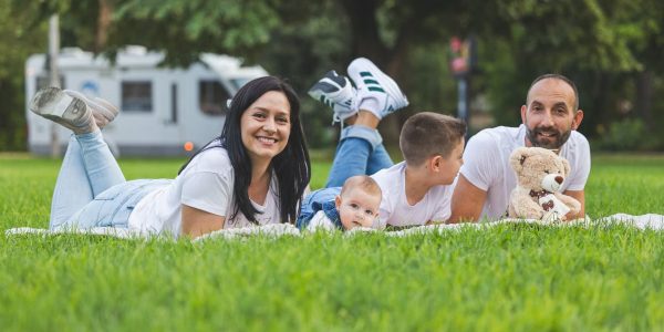 Happy family lying on grass with RV in the background, enjoying a sunny outdoor picnic.
